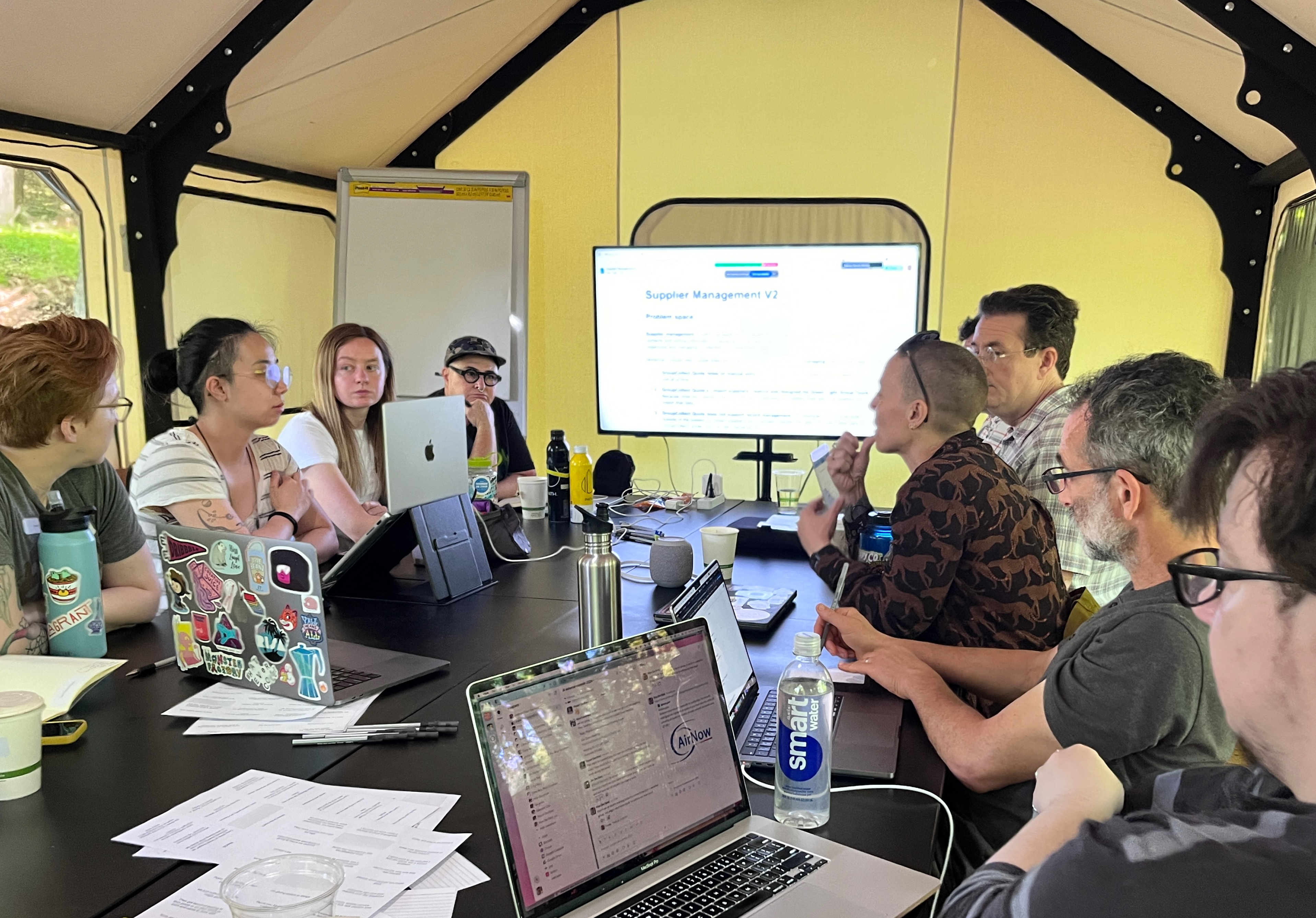 A group of Flagrant team members are seated at a table covered in pens and paper, watching another team member standing next to a wall with a big piece of paper covered in bright sticky notes. Everone's attention is on the sticky notes.