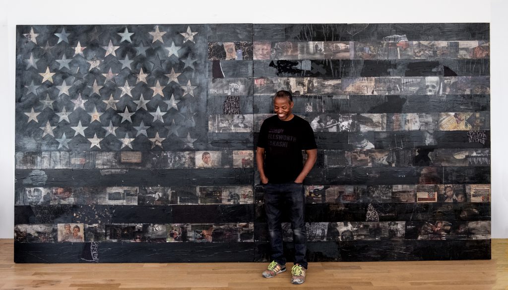 Man looking down standing in front of a large collage of a black and brown American flag. 