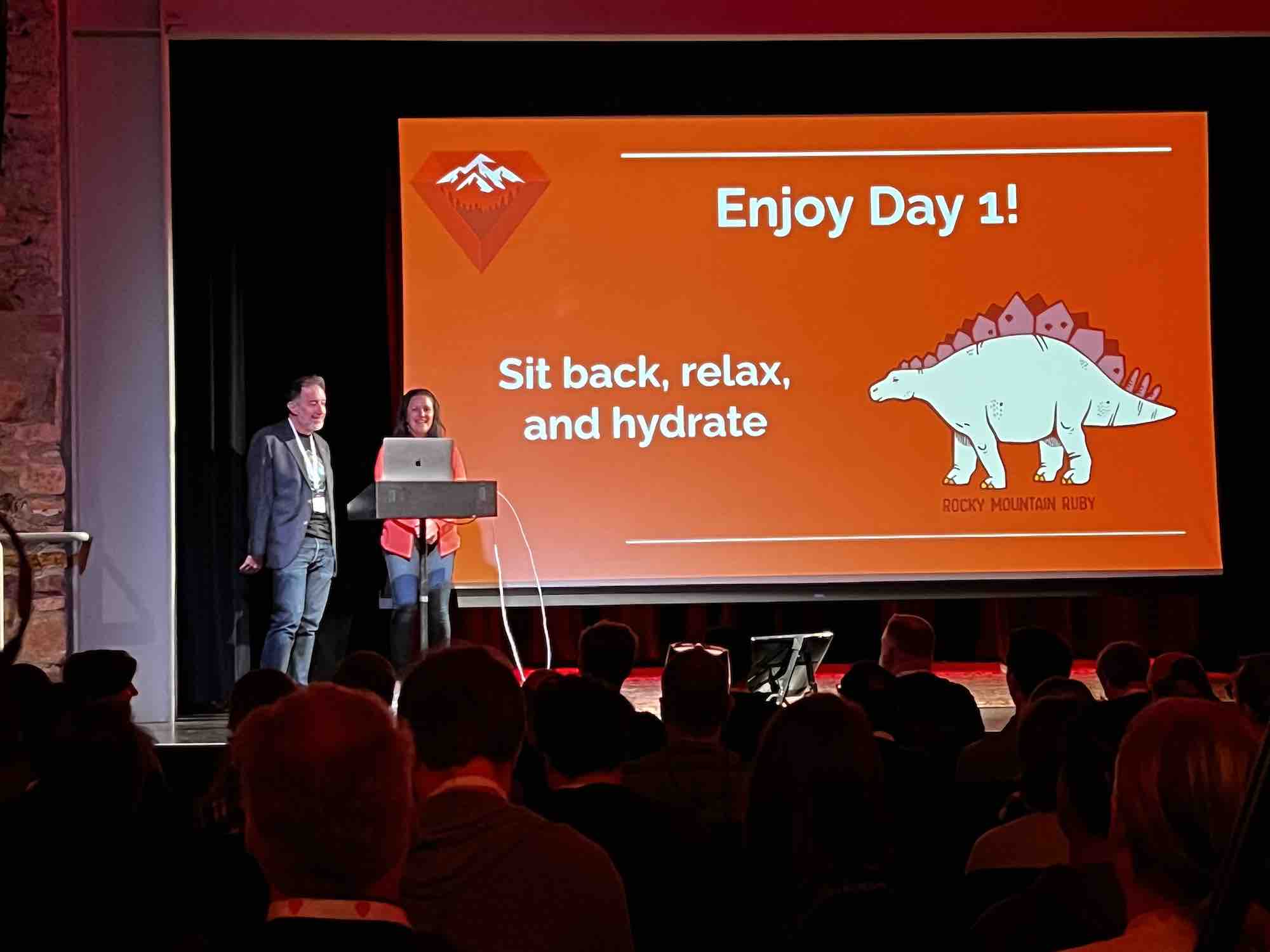 Two speakers on stage at Rocky Mountain Ruby conference stand in front of a large screen with a message: 'Enjoy Day 1! Sit back, relax, and hydrate.' The slide features an illustrated stegosaurus